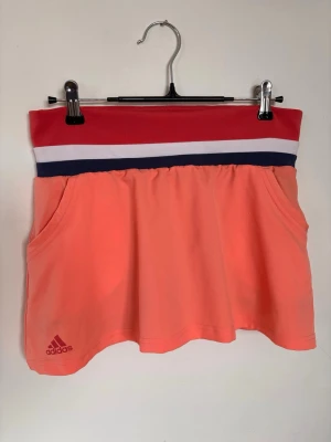 Adidas korall padelkjol med bollfickor - Sportig skort från Adidas i korallorange funktionsmaterial. Bred midjeresår i korall med vita och marinblå ränder. Två fickor framtill och diskret Adidas-logga. Perfekt för träning eller tennis, ger både rörelsefrihet och stil. Även en bollficka på var sida av innershorts!