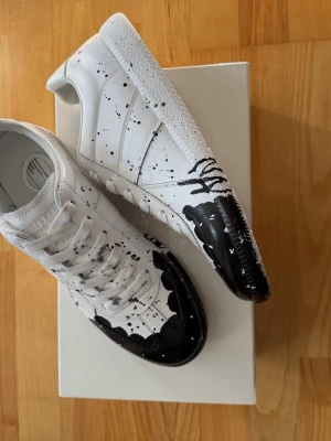 Maison Margiela vit med svart  - Maison Margiela sneakers med svart drip och färgstänk-design på tå och sidor. Skorna har klassisk låg profil, vita snören och sula. Tillverkade i slätt läder eller konstläder för en clean look med edgy detaljer.