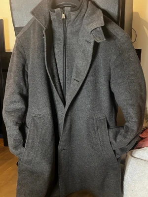 Mörkgrå kashmirrockfrån Hugo boss  - Stilren mörkgrå kashmirrock från Hugo BOSS med klassisk krage och dold dragkedja samt knäppning. Rocken har två sidofickor och en innerficka, samt ett avtagbart innerfoder med hög krage för extra värme. Perfekt för kyliga dagar och ger en clean look. Nypris 5500:- mitt pris 1800:- 