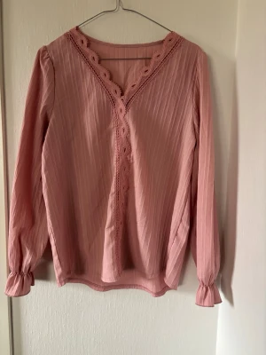 Rosa blus med spetsdetaljer - Söt rosa blus med långa ärmar och v-ringning dekorerad med spets. Blusen har diskreta vertikala ränder och lätt puffade ärmslut med resår. Perfekt för en trendig och feminin look.