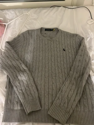 Grå kabelstickad tröja Polo Ralph Lauren - Snygg grå kabelstickad tröja från Polo Ralph Lauren med klassisk rund halsringning och broderad logga på bröstet. Tröjan har långa ärmar och ribbade muddar vid ärmslut och nederkant. Perfekt för lager på lager och stilren look. Storlek S men passar M mer