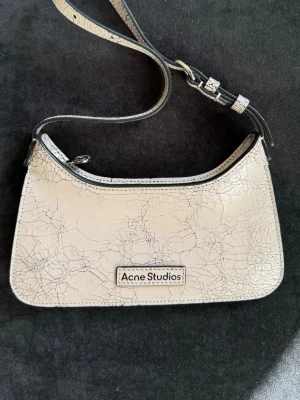 Light Pink handväska Acne Studios - Snygg vit handväska från Acne Studios i ett coolt krackelerat läder. Väskan har en justerbar axelrem och en unik detalj med spegel och knapp på baksidan. Perfekt för dig som gillar trendiga accessoarer med en twist.