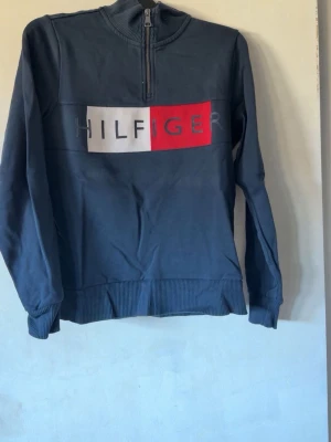 Blå half-zip hoodie Tommy Hilfiger - Snygg blå hoodie från Tommy Hilfiger med half-zip och stor logga framtill i vitt och rött. Tröjan har ribbade muddar vid ärmslut och nederkant samt hög krage. Perfekt för en avslappnad och trendig look.