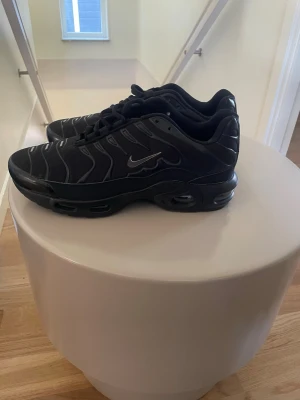 Nike Air Max Plus svarta sneakers - Nike Air Max Plus sneakers i helsvart med reflekterande detaljer och ikonisk Swoosh på sidan. Skorna har en vågig design på ovandelen, synliga Air-enheter i sulan och är tillverkade i en mix av mesh och syntetmaterial. Perfekta för dig som gillar streetwear.