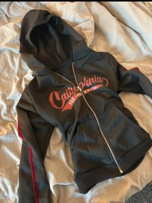 Svart hoodie med röd text California - •Säljer denna svarta hoodie med dragkedja och huva. Två fickor framtill och ribbade muddar. Tillverkad i mjukt och bekvämt material!                                                                   Märke• Shein                                                        Köpt för• Ca 200kr                                             Mitt pris• 100kr                                                    Storlek• M