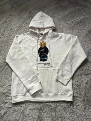 Vit hoodie Polo Ralph Lauren björn - Vit hoodie från Polo Ralph Lauren med ikonisk björn-tryck på bröstet. Hoodien har huva med dragsko och känguruficka framtill. Tillverkad i en mjuk bomullsblandning för skön känsla. Perfekt för dig som gillar klassisk streetwear med en twist.