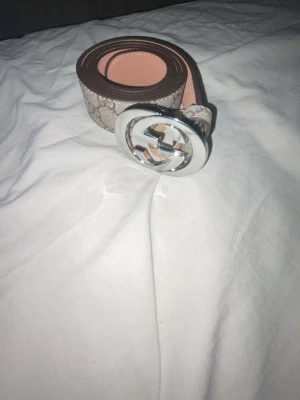 GG bälte - Säljer ett snyggt beige bälte från GG med klassiskt GG-mönster och ett stort, rundat silverfärgat spänne med logotyp. Bältet är tillverkat i skinn och har en stilren design som passar till många outfits.