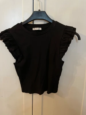 Svart volangärmad topp från Zara - Snygg svart topp från Zara med volangdetaljer vid ärmhålen. Toppen är varsamt använd då den ändast blivit använd vid ”fina” tillfällen. Super fin på men säljs då jag växt ur den, jag skulle säga att toppen passar perfekt till xxs-m, själv brukar jag använda S på tröjor men tycker att denna blivit lite kort då jag växt! Skulle nog säga att modellens storlek M är som en vanlig tröja i xs/s men den är väldigt stretchig så den bör passa M också!