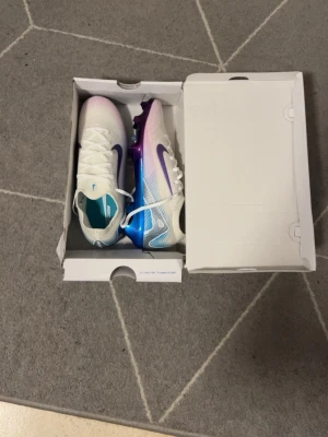 Nike vita/lila Mercurial fotbollsskor - Säljer ett par Nike Mercurial fotbollsskor i vitt med lila och blå detaljer. Skorna har en snygg gradient från vitt till lila och blått, samt en lila Nike swoosh på sidan. Tillverkade i syntetmaterial för bra grepp och lätt känsla på planen. Perfekta för dig som vill sticka ut på matchen.