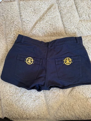 Mörkblå shorts med guldiga detaljer - Snygga mörkblå shorts med låg midja och två bakfickor som pryds av guldiga TEAM-emblem. Klassisk passform och bälteshällor upptill. Perfekta för sommarens alla äventyr.