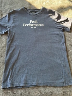 Blå Peak Performance t-shirt - Snygg blå t-shirt från Peak Performance med vit logga och text 'Est. 1986' på bröstet. Klassisk rund hals och korta ärmar. Tillverkad i mjuk bomull för skön känsla och enkel stil. Ny pris 499