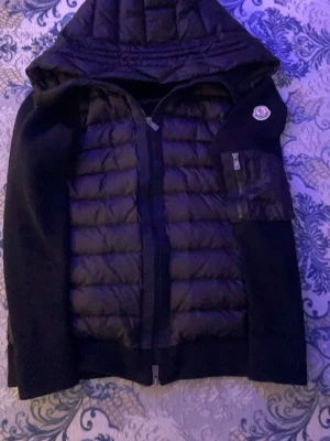 Svart pufferjacka med huva från Moncler - Snygg svart pufferjacka från Moncler med huva och quiltad front. Jackan har dragkedja framtill, en ficka med dragkedja på ärmen och Moncler-logga på vänster ärm. Materialmix med vadderad kropp och stickade ärmar ger en modern look.