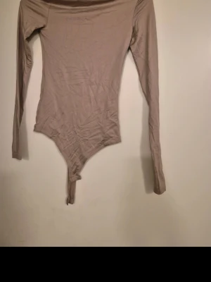 Beige långärmad body - Säljer en enkel och stilren beige body med långa ärmar. Bodyn har en rund halsringning och är tillverkad i ett mjukt, stretchigt material som sitter tajt mot kroppen. Perfekt att matcha med jeans eller kjol för en clean look.