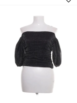  topp - Trendig svart offshoulder topp med glittrigt tyg och puffiga trekvartsärmar. Sitter snyggt figurnära över kroppen och har en croppad passform som passar perfekt till höga jeans eller kjol. Perfekt för dig som vill sticka ut lite extra.