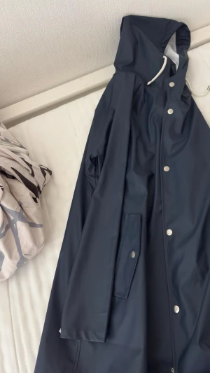 Mörkblå regnjacka med huva 157 - Snygg mörkblå regnjacka från 157 med huva och vita dragsnören. Jackan har tryckknappar framtill, fickor med lock och är tillverkad i ett vattenavvisande material. Perfekt för regniga dagar och enkel att matcha med olika outfits.