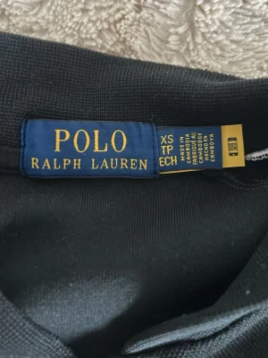 Svart hoodie från Polo Ralph Lauren - Svart hoodie från Polo Ralph Lauren med dragkedja framtill och två fickor. Klassisk röd Polo-logga broderad på bröstet och vita detaljer på axlarna. Tillverkad i mjukt material som känns skönt mot huden. Perfekt för en avslappnad och stilren look.