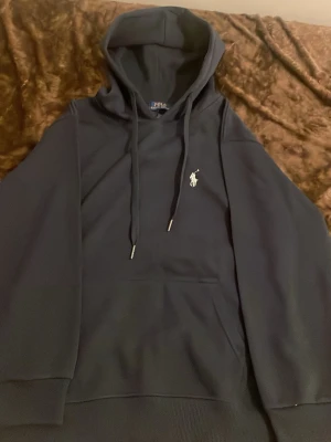 Mörkblå hoodie från Polo Ralph Lauren - Snygg mörkblå hoodie från Polo Ralph Lauren med klassisk vit broderad logga på bröstet. Hoodien har dragsko i huvan och en stor magficka framtill. Tillverkad i mjukt bomullsmaterial som känns riktigt skönt mot huden. Perfekt för en avslappnad och stilren look.