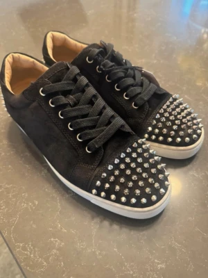 Christian Loubotin sneakers  - Säljer ett par svarta sneakers från Christian Louboutin med coola silverfärgade nitar på tå och häl. Skorna har svart mocka, svarta skosnören och vit sula. Insidan är beige och de har en riktigt edgy vibe. Perfekta för dig som vill sticka ut med din stil.