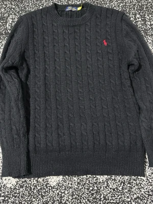 Svart kabelstickad tröja Polo Ralph Lauren - Kabelstickad svart tröja från Polo Ralph Lauren med rund halsringning och röd broderad logga på bröstet. Tröjan har långa ärmar och ribbade muddar vid ärmslut och nederkant. Klassisk och tidlös design i mjukt material.