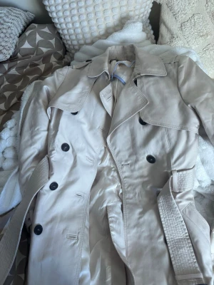 Beige trenchcoat  - Stilren beige trenchcoat från Zara Woman med klassisk dubbelknäppning, bred krage och bälte i midjan. Jackan har svarta knappar och axelklaffar för en tidlös look. Perfekt för dig som vill ha en snygg och enkel trenchcoat till garderoben.