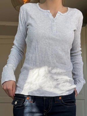 Vit ribbad långärmad topp - Långärmad tröja från Brandy Melville i vit/ljusgrå ribbad bomull med knappdetaljer framtill. Sitter snyggt och har en enkel, clean look som passar till det mesta. Endast använd 1 gång, som ny. Vid frågor eller önskemål om fler bilder är det bara att skriva privat 💕
