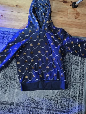 Blå hoodie från Snipes x McDonald's - Cool blå hoodie med McDonald's logga i gult och vitt geometriskt mönster över hela plagget. Samarbete mellan Snipes och McDonald's, med stor huva och ribbade muddar. Perfekt för dig som gillar streetwear och unika designer.