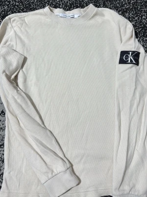 Vit långärmad tröja Calvin Klein - Vit långärmad tröja från Calvin Klein Jeans med ribbade muddar och ett stort svart CK-märke på ena ärmen. Tillverkad i ett strukturerat, lätt ribbat material som ger en avslappnad men ändå stilren look.