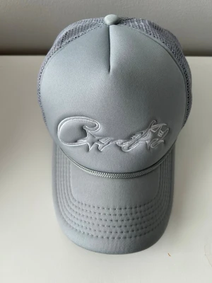 Corteiz keps - Snygg grå truckerkeps från Crude med broderad logga framtill. Kepsen har mesh baktill för extra ventilation och justerbar snapback. Perfekt för en avslappnad streetwear-look och passar till det mesta.