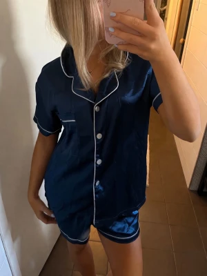 Mörkblå satinpyjamas med vita detaljer - Snygg mörkblå pyjamas i glansigt satinmaterial med kortärmad skjorta och matchande shorts. Setet har vita kontrastsömmar och knappar framtill samt en bröstficka. 
