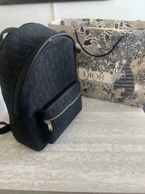 DlOR BACKPACK - Säljer nu min DlOR Rider Backpack i det klassiska svarta Oblique jacquard-mönstret. * Skick: Nyskick * Material: Slitstark canvas med detaljer i svart kalvskinn. * Detaljer: Tvåvägsdragkedja, stort huvudfack invändigt samt en praktisk framficka. Justerbara vadderade axelremmar för hög komfort. * Mått: Ca 30 x 42 x 15 cm. * Nypris: Ca 24 000 – 26 000 SEK. * Medföljer: Original dustbag  Autenticitet garanteras. Skickas spårbart och försäkrat. Tveka inte att höra av dig vid frågo