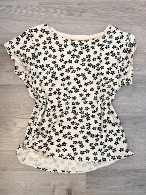 Blommig topp från Holly & Whyte - Söt topp från Holly & Whyte by Lindex i vitt med svart blommigt mönster. Modellen har korta ärmar och en lätt figurnära passform. Perfekt till jeans eller kjol för en fräsch look.