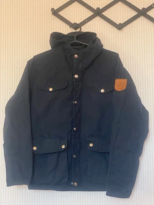 Marinblå vindjacka från Fjällräven - Snygg marinblå vindjacka från Fjällräven med huva, fyra framfickor med lock och tryckknappar samt klassisk läderpatch på ärmen. Jackan har en rak passform och stängs med dragkedja och tryckknappar. Materialet är slitstarkt och passar perfekt för uteliv.