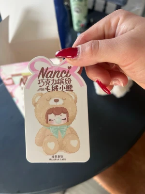 Nanci Hazelnut Latte nalle - Söt mjukisdjur från Nanci, modell Hazelnut Latte. Nallen är ljusbrun med ett mintgrönt band runt halsen och har hjärtformade detaljer på tassarna. Perfekt som dekoration eller present till någon som gillar gulliga samlarfigurer.