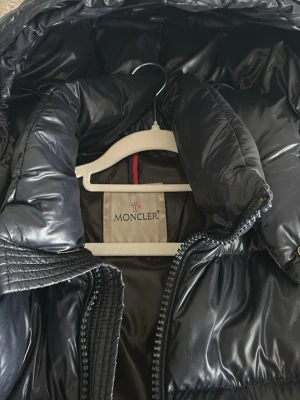 Svart dunjacka från Moncler - Snygg svart dunjacka från Moncler med glansig finish och quiltad design. Jackan har hög krage, avtagbar huva och två dragkedjeförsedda fickor framtill. Perfekt för kalla vinterdagar och riktigt trendig look.