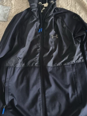 Svart vindjacka från Under Armour - Snygg svart vindjacka från Under Armour med dragkedja framtill, huva med snörning och blå detaljer. Jackan har logga på bröstet och ärmen samt praktiska fickor med dragkedja. Perfekt för dig som gillar sportig stil.