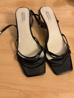 Svarta sandaler med remmar från NLY Shoes - Säljer ett par svarta sandaler från NLY Shoes med tunna, glansiga remmar och fyrkantig tå. De har en stilren och trendig look med justerbar rem runt hälen. Materialet är lackat för en extra snygg finish.