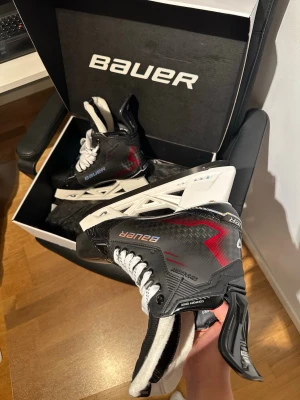 Bauer skridskor svart/röd - Säljer ett par snygga Bauer skridskor i svart med röda detaljer och vita snören. Skridskorna har en robust och sportig design med vita skenor och är perfekta för hockey eller skridskoåkning. Kommer i originalkartong.