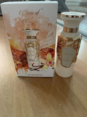 Fragrance World Crème of Clouds Eau de Parfum - Lyxig parfymflaska i vitt med gulddetaljer och elegant mönster. Flaskan har en exklusiv design och kommer i en snygg förpackning med illustrationer av karamell, vanilj och kanelbulle. Perfekt för dig som gillar söta och varma dofter. Endast testad!