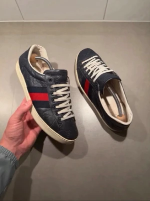 Gucci blå sneakers med röd rand - Snygga mörkblå sneakers från Gucci med klassisk röd och marinblå rand på sidan. Skorna har vita snören, vit sula och är tillverkade i präglat läder med diskret GG-mönster. Perfekta för dig som vill ha en stilren och exklusiv look.