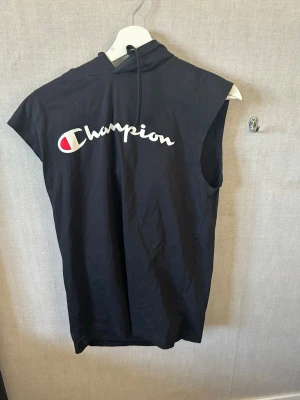 Svart ärmlös topp från Champion - Svart ärmlös topp från Champion med stor vit logga framtill och huva med dragsko. Perfekt för en sportig look och enkel att matcha. Tillverkad i mjuk bomull för skön känsla.