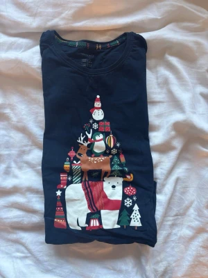 Mörkblå julpyjamaströja Esmara - Mörkblå långärmad pyjamaströja från Esmara med juligt tryck framtill. Motivet visar en isbjörn, ren, pingvin, snögubbe och julklappar i färgglada toner. Rund halsringning och mjukt bomullsmaterial gör den perfekt för mysiga vinterkvällar.