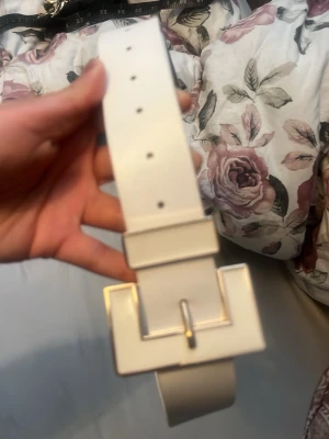 Vitt bälte med fyrkantigt spänne - Stilrent vitt bälte med bred rem och ett stort, fyrkantigt spänne i silverfärg. Perfekt accessoar för att lyfta din outfit med en modern touch. Bältet har flera hål för justerbar passform och är tillverkat i syntetmaterial.