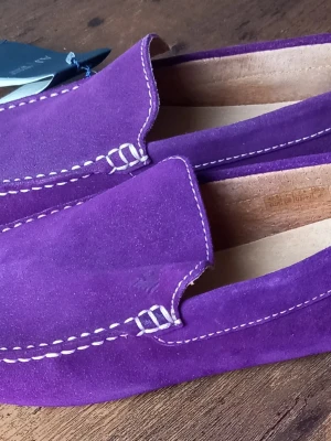 Lila mockaloafers med vit söm från Armani Jeans  - Säljer ett par loafers från Armani Jeans i lila mocka med kontrasterande vita sömmar. Skorna har en klassisk, stilren design med fyrkantig tå och mjukt innermaterial. Perfekta för dig som vill sticka ut med färg och ändå hålla det snyggt och enkelt.