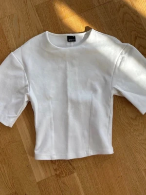 Vit kortärmad topp från Gina Tricot - En stilren vit topp från Gina Tricot med korta ärmar och rund halsringning. Modellen är något croppad och har diskreta sömdetaljer framtill som ger en snygg passform. Perfekt för att skapa en clean look.