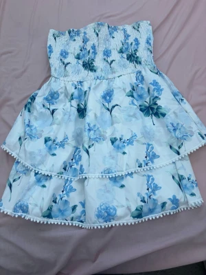 Vit blommig volangkjol med blå toner - Supersöt kort kjol med blått blommönster på vit botten. Kjolen har smock i midjan och flera lager med volanger, samt små bollar längs kanterna för extra detalj. Perfekt för sommarens alla tillfällen!