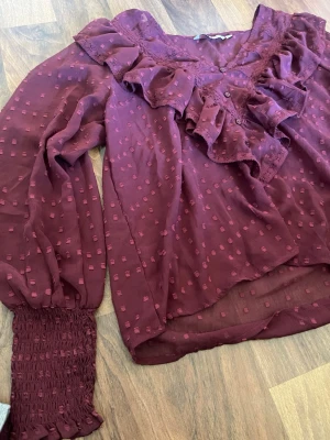 Vinröd blus med volang från ONLY - Vinröd blus från ONLY med långa ärmar, volangdetaljer längs v-ringningen och diskret strukturmönster. Blusen har smockade ärmslut och är tillverkad i ett lätt, transparent tyg som ger en luftig känsla, använd typ 3 gånger.