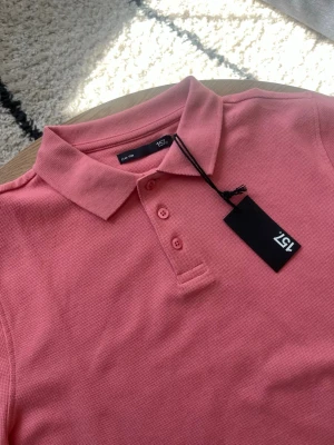 Rosa piké från 157 - Tjena👋 Säljer denna snygga rosa pikétröja från 157 med klassisk krage och knappar framtill👌 Tillverkad i mjuk bomull som känns skön mot huden🤩 Perfekt till jeans eller shorts för en avslappnad look✌️ Meddela gärna vid frågor📩