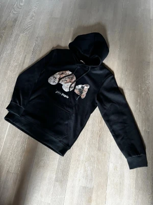 Svart hoodie från Hype Angels - Svart hoodie från Hype Angels med stor huva och magficka. Framsidan har ett stort tryck i form av bokstäver med mönster och texten 'Hype Angels' under. Mjuk bomullskänsla och ribbade muddar vid ärmslut och nederkant. Storlek s men sitter mer som xs