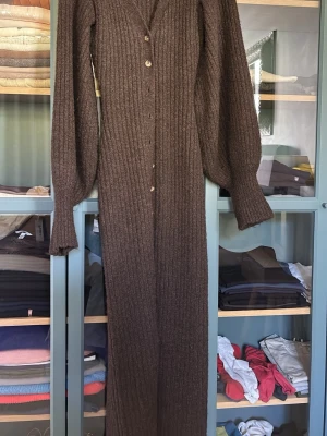 Brun lång stickad klänning - Lång, ribbstickad brun klänning med v-ringning och knäppning framtill. Klänningen har vida ärmar med markerade muddar och är tillverkad i ett mjukt, värmande material. Perfekt för lager-på-lager och en snygg höstlook.