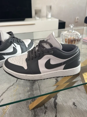 Nike Air Jordan 1 Low grå/vit - Snygga Nike Air Jordan 1 Low sneakers i svart och vitt läder. Klassisk siluett med perforerad tåbox, grå swoosh och gråa skosnören. Perfekta för dig som gillar streetwear och vill ha en ikonisk look.